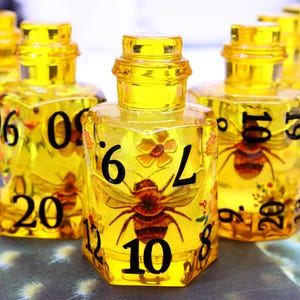 May include: Yellow, hexagonal dice-shaped bottles with black numbers and bee illustrations inside. The bottles have a clear, glass-like appearance with a yellow tint. The numbers are large and easy to read.