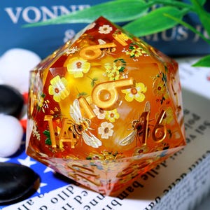 May include: A large, multi-sided die with a transparent amber resin body. The die features gold-colored numbers and is decorated with yellow flowers and detailed bee illustrations. The number 14 and 16 are visible.