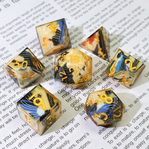 May include: A set of seven polyhedral dice with a cream base, gold numerals, and blue and black abstract patterns. These dice are for tabletop games and are displayed on a printed paper background.