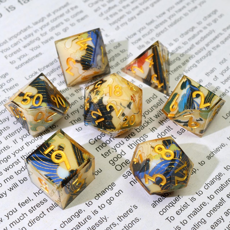 May include: A set of seven polyhedral dice with a cream base, gold numerals, and blue and black abstract patterns. These dice are for tabletop games and are displayed on a printed paper background.