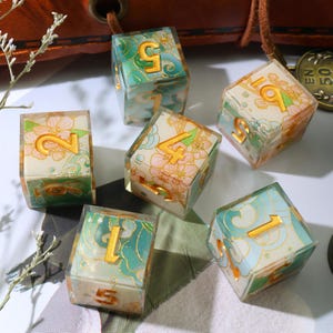 May include: Set of six clear, cube-shaped dice with gold numbers and decorative floral and wave patterns in shades of green, blue, and pink. The dice are designed for tabletop games.