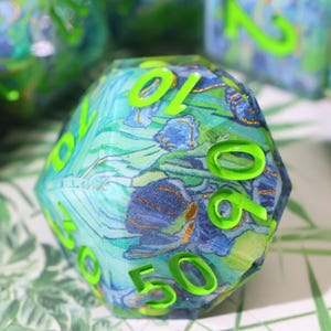 May include: A multi-faceted die featuring a blue and green floral design with gold accents. The numbers are a bright neon green. The die has a clear, glossy finish, and appears to be made of resin. The die is a unique gaming accessory.