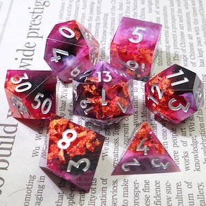 Peut inclure: Ensemble de dés polyédriques avec un design vibrant et superposé. Les dés présentent un mélange de rouge profond, de violet et d'accents de feuilles d'or, avec des chiffres argentés. Idéal pour les jeux de société.