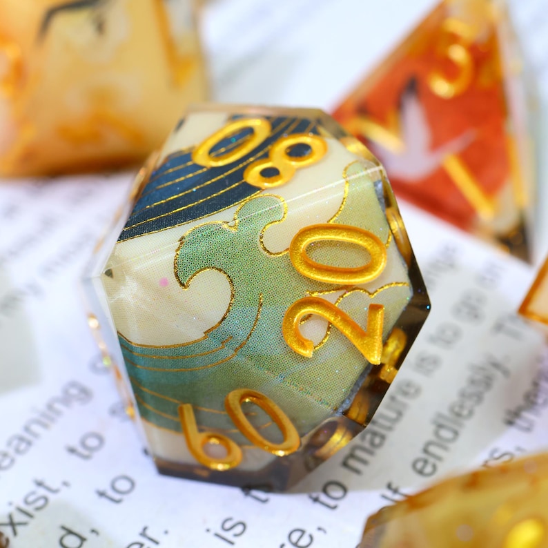 May include: A clear, multi-sided die with gold numbering and a Japanese wave pattern in blue and green. The die has a gold border. The background is blurred with other dice.