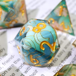 May include: Close-up of a set of polyhedral dice with a blue and teal wave pattern and gold numbering. The dice are made of a clear resin material. The numbers are in a gold color. The dice are for tabletop games.