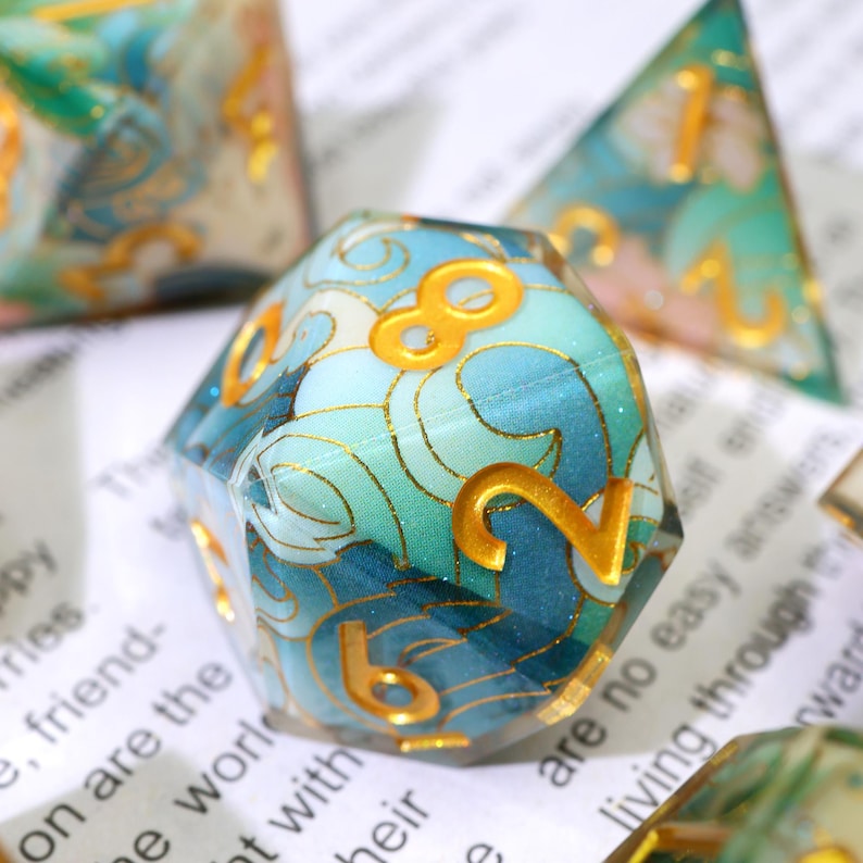 May include: Close-up of a set of polyhedral dice with a blue and teal wave pattern and gold numbering. The dice are made of a clear resin material. The numbers are in a gold color. The dice are for tabletop games.