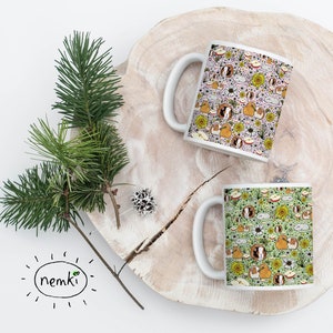 May include: Two white ceramic mugs with a repeating pattern of guinea pigs, sunflowers, and apples. One mug has a light purple background, and the other has a light green background. The mugs are on a wooden surface with a sprig of pine.