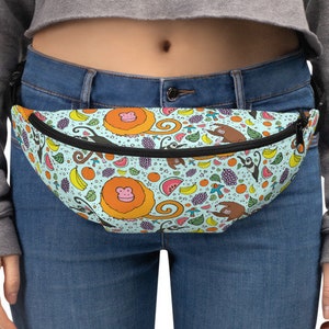 May include: A light blue fanny pack with a colorful pattern of monkeys, fruit, and swirls. The fanny pack has a black zipper and strap. The bag is worn around the waist.