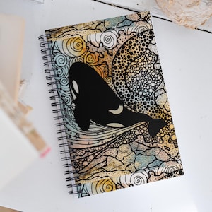 May include: Spiral-bound notebook featuring a black and white orca illustration. The cover design includes a detailed, colorful background with swirls, dots, and lines in shades of yellow, blue, and brown. The notebook is open, revealing the spiral binding.
