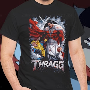 May include: Black t-shirt featuring a graphic of a superhero in a red and white costume, with the word "THRAGG" in a distressed font. The design includes a cape and a battle scene. The background shows a character's face.