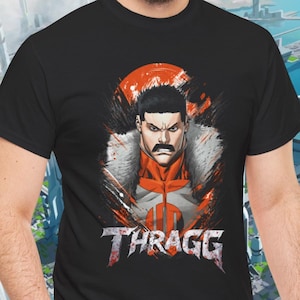 May include: Black t-shirt featuring a graphic of a stern-faced character with a mustache, wearing a red and white outfit with a fur-lined collar. The word "THRAGG" is displayed in a stylized font below the character.