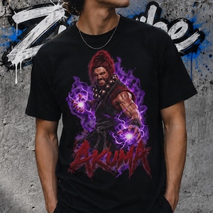 Può includere: Una t-shirt nera a maniche corte con una stampa grafica del personaggio Akuma circondato da energia viola. Il personaggio ha i capelli rossi, indossa abiti scuri con perline e tiene in mano sfere viola luminose. Il nome Akuma è stampato in rosso.