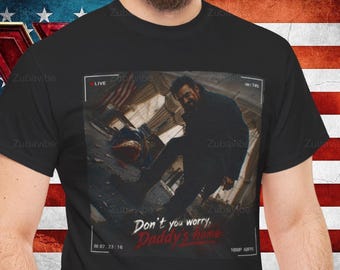 May include: Black t-shirt featuring a graphic of a man in a dark setting with the text "Don't you worry, Daddy's home." The image has a "LIVE" overlay and a timestamp. The shirt is set against an American flag.