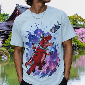 Puede incluir: Una camiseta azul claro con un estampado gráfico de un dinosaurio rojo que lleva un collar con la palabra "DEVIL" impresa, situado frente a un horizonte urbano estilizado, con una pequeña orca nadando en el aire sobre el dinosaurio.