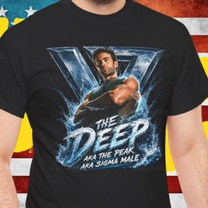Könnte beinhalten: Schwarzes T-Shirt mit einer Grafik eines Mannes und dem Text "THE DEEP" und "aka the peak aka sigma male" in Weiß und Blau. Das Design beinhaltet Wassereffekte und eine geometrische Form.