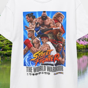 Puede incluir: Una camiseta blanca de manga corta con un estampado gráfico colorido de personajes de Street Fighter como Ryu, Chun-Li y Zangief. El texto dice Street Fighter The World Warrior con pequeñas banderas nacionales y el logo de Capcom.