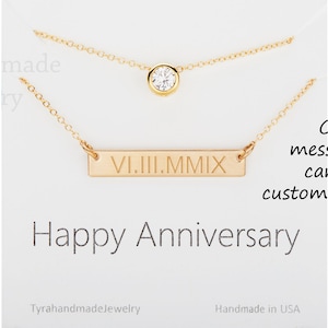 May include: Two gold necklaces on a white card. The top necklace has a single clear gemstone. The bottom necklace has a rectangular bar with Roman numerals. The card reads "Happy Anniversary".