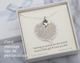 Puede incluir: Un collar con colgante de hoja plateada presentado en una caja de regalo blanca con una cinta azul claro. La tarjeta interior dice: "Getting so excited for the arrival of your little girl." También se ve el texto "Card message can be personalized".