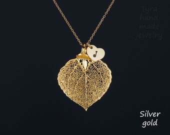 Puede incluir: Collar dorado con un delicado colgante de hoja y un pequeño dije de corazón con la letra "J". El colgante de hoja muestra intrincados patrones de venas. El collar está sobre un fondo oscuro. El texto "Tyra hand made jewelry" está en el fondo.