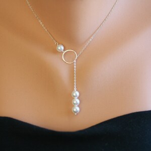 Puede incluir: Un delicado collar de plata con un pequeño colgante circular y tres perlas colgantes. Una sola perla está colocada a un lado del círculo. El collar se muestra en una persona.