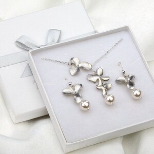 May include: A white gift box with a silver ribbon contains a silver necklace and earrings set. The necklace features a delicate chain with a pendant of three silver orchid flowers. The earrings are silver orchid flowers with a pearl drop.