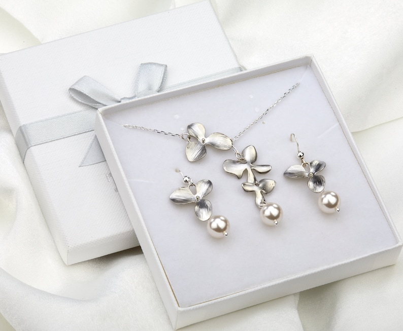 May include: A white gift box with a silver ribbon contains a silver necklace and earrings set. The necklace features a delicate chain with a pendant of three silver orchid flowers. The earrings are silver orchid flowers with a pearl drop.