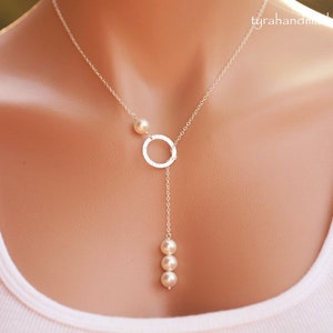 May include: A delicate silver necklace with a hammered circle pendant and a vertical drop of three small pearls. A single pearl accents the chain. The necklace is worn against a white top.