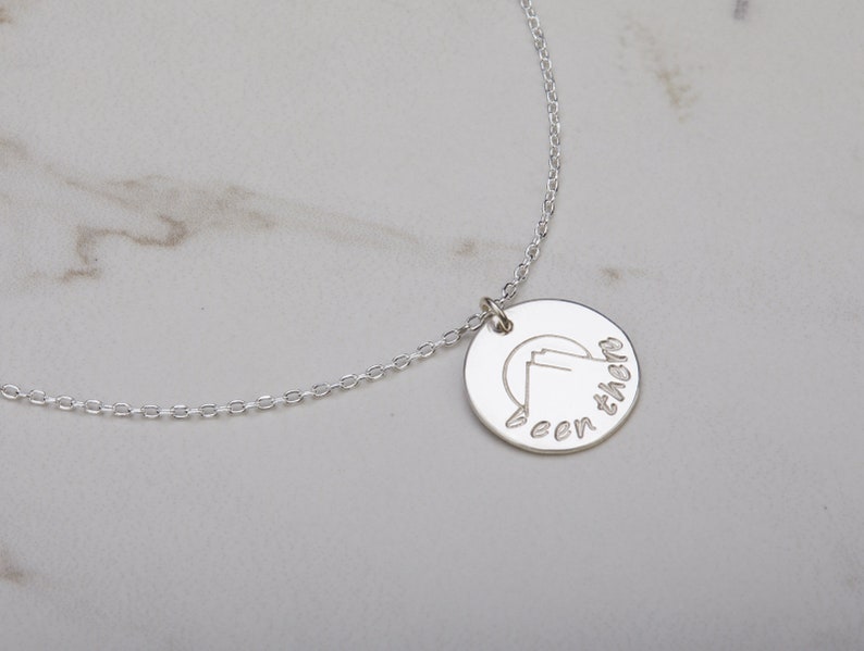 May include: A silver necklace with a round pendant that has a mountain range silhouette and the text "been there" engraved on it.