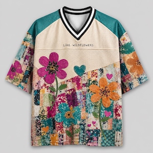 May include: A short-sleeve V-neck jersey featuring a cream-colored upper section with the text LIKE WILDFLOWERS, and a lower section with a colorful patchwork pattern. The shirt is decorated with large pink, orange, and teal floral appliqués and hearts.
