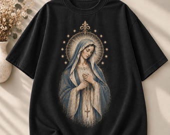 May include: A black short-sleeve t-shirt featuring a central graphic of a figure wearing a blue and white head covering and robe, holding a rosary with a cross. The figure is surrounded by a golden halo with star accents and a fleur-de-lis.