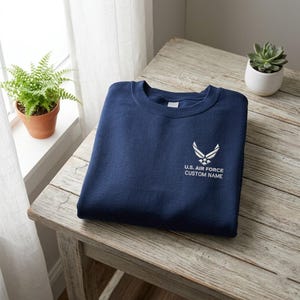 Könnte beinhalten: Ein marineblaues Sweatshirt mit dem Logo der US Air Force und dem Text "CUSTOM NAME" in Weiß. Das Sweatshirt liegt gefaltet auf einem rustikalen Holztisch, im Hintergrund eine kleine Topfpflanze.