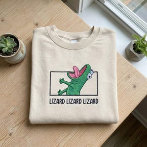 May include: Beige sweatshirt with an embroidered green lizard design inside a black rectangle. The lizard has its tongue sticking out and the words "LIZARD LIZARD LIZARD" are embroidered below the design. The sweatshirt is folded on a wooden surface.