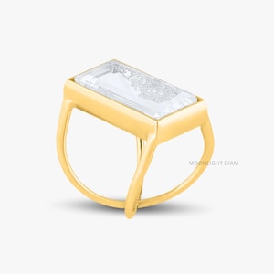 May include: A gold ring with a rectangular, clear gemstone. The ring has a distinctive open band design with curved lines. The gemstone is set in a gold bezel, giving it a modern and elegant appearance. The ring is shown against a white backdrop.