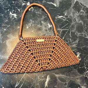 May include: A trapezoidal handbag crafted from metallic bronze yarn in a dense crochet stitch. The bag features a single rounded handle and a small rectangular gold-toned metal plaque centred on the top edge, showcasing a structured design.