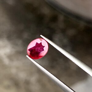 May include: A faceted, oval-shaped ruby gemstone held by tweezers. The ruby is a deep red color with a star-like pattern visible within the stone. The tweezers are silver-colored, and the background is blurred.