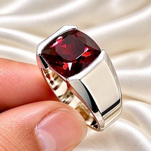Peut inclure: Une bague en métal argenté poli dotée d'une grande pierre précieuse rouge foncé de taille carrée, montée sur un anneau large et lisse. Le sertissage met en valeur la surface facettée de la pierre rouge, contrastant avec le fini métallique brillant.