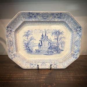 May include: An octagonal ceramic platter featuring a blue and white transferware pattern. The center depicts a landscape with a building, trees, and a small boat on water. The wide border displays intricate floral and scrollwork motifs.