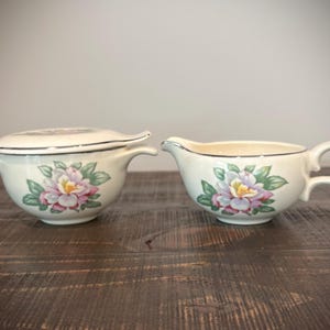 May include: A cream-colored ceramic sugar bowl with a matching lid and a matching creamer pitcher. Both pieces feature a floral pattern with pink and purple petals and green leaves, accented by a thin silver rim along the top edges.