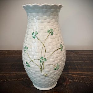 May include: A white ceramic vase with a textured basket-weave pattern and a scalloped rim. The surface is decorated with a hand-painted green shamrock motif featuring delicate stems and leaves extending upward across the front of the vessel.