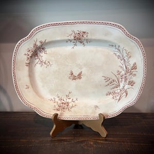 May include: An oval ceramic platter featuring a cream-colored base with brown botanical illustrations of flowers and butterflies. The rim is decorated with a repeating brown pattern, and the center displays a butterfly motif.