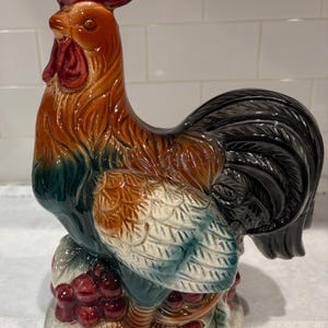 May include: A ceramic rooster figurine with a red comb and wattle, brown and orange body, and black tail feathers. The rooster is perched on a base with red grapes and green leaves. The figurine is a decorative item.