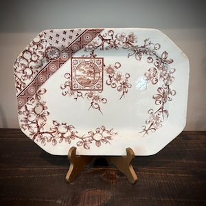 May include: An octagonal ceramic serving platter featuring a white base with brown transferware patterns. The design includes a central square landscape medallion, floral branches, and a geometric border pattern along the upper left corner.
