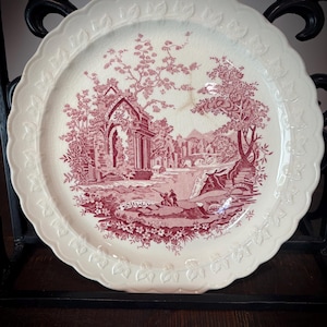 May include: A cream-colored ceramic plate with a scalloped edge featuring a raised leaf pattern. The center displays a detailed maroon transferware print of a landscape with stone ruins, trees, and a small figure near a body of water.