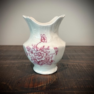 May include: A white ceramic pitcher featuring a scalloped rim and a flared base. The body of the pitcher is decorated with a detailed magenta floral transfer print, showcasing a large central flower surrounded by smaller blossoms and foliage.