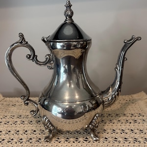 May include: A silver-colored tea or coffee pot with a decorative handle and spout. The pot has a lid with a finial and sits on three small feet. The pot has an ornate design and a reflective surface.