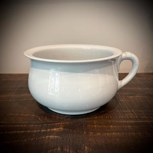 May include: A white ceramic chamber pot with a smooth, glossy finish, a wide circular rim, and a single curved handle attached to the side. The vessel features a rounded, deep basin design with a stable base, presented in a clean, solid white color.