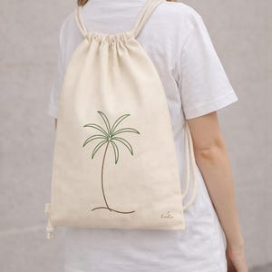 Puede incluir: Mochila con cordón de color crema con un diseño minimalista de palmera en verde y marrón. La bolsa tiene una estética sencilla y limpia con cierre de cordón y correas de cuerda. La bolsa está hecha de una tela suave y natural.