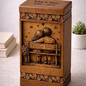 Puede incluir: Urna de madera con dise&ntilde;o tallado. La parte superior est&aacute; grabada con "John & Mary 1985-2024". La parte frontal presenta una pareja en un banco con las palabras "Forever Love". La urna es marr&oacute;n con detalles de hojas y flores.