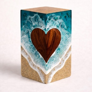 May include: A wooden block with a heart-shaped cutout, featuring a beach scene design. The top is wood, the sides have a turquoise resin ocean design with white waves, and a sandy base. The heart is made of wood.
