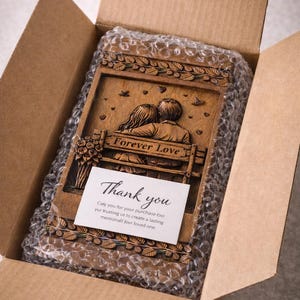 Puede incluir: Placa de madera con las palabras "Forever Love" y una pareja en un banco, dentro de una caja de cart&oacute;n. La placa tiene un dise&ntilde;o floral y una tarjeta que dice "Gracias". La placa est&aacute; envuelta en pl&aacute;stico de burbujas.